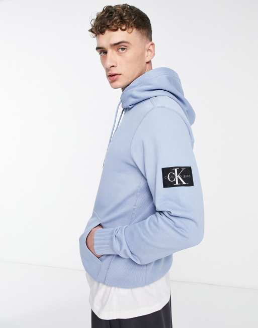 Calvin Klein Jeans – Jasnoniebieska bluza z kapturem i czarno-białą naszywką z logo na rękawie