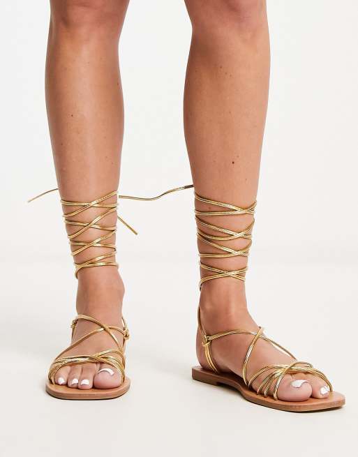 London Rebel – Guldmetalliska platta sandaler med knytning runt benet