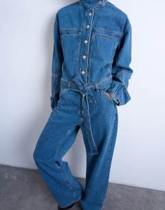 Model wearing Denim funnel neck belted jumpsuit in mid blue